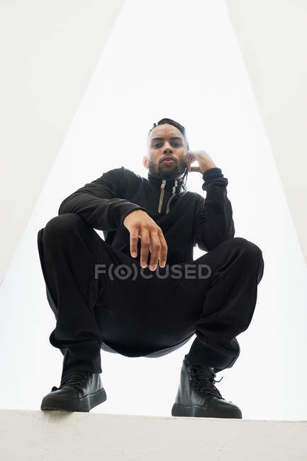 African American man in black clothes with braided hair crouching on white background — Stock Photo