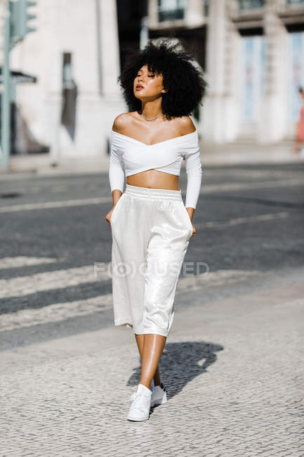Mulher afro-americana em branco roupa de moda em pé com as mãos em bolsos na beira da estrada contra o fundo urbano — Fotografia de Stock