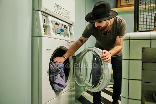 Joven barbudo guapo hombre en sombrero poniendo cosas en la lavadora en la sala de lavandería - foto de stock