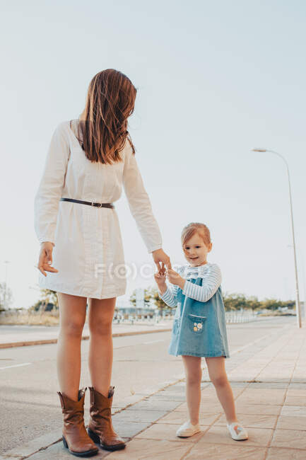 Anonymous young woman and little girl holding hands and standing on sidewalk together against cloudless blue sky on city street — Stock Photo