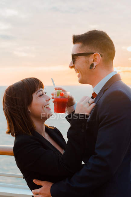 Side view of young attractive couple drinking red beverage with straws from one glass on background of sunset sea — Stock Photo