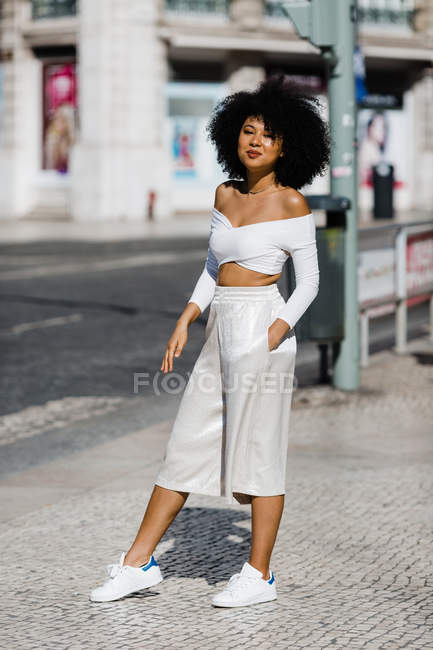 Lächelnde Afroamerikanerin im weißen modischen Outfit, die mit der Hand in der Tasche am Straßenrand vor urbanem Hintergrund steht — Stockfoto