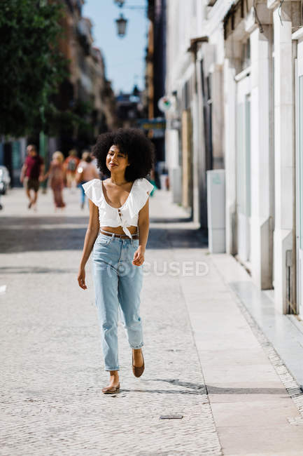 Attraente donna alla moda in jeans e crop top godendo di camminare sulla giornata estiva su sfondo urbano sfocato — Foto stock