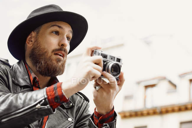 Joven hombre guapo barbudo en sombrero negro y chaqueta de cuero tomando fotos en la calle - foto de stock