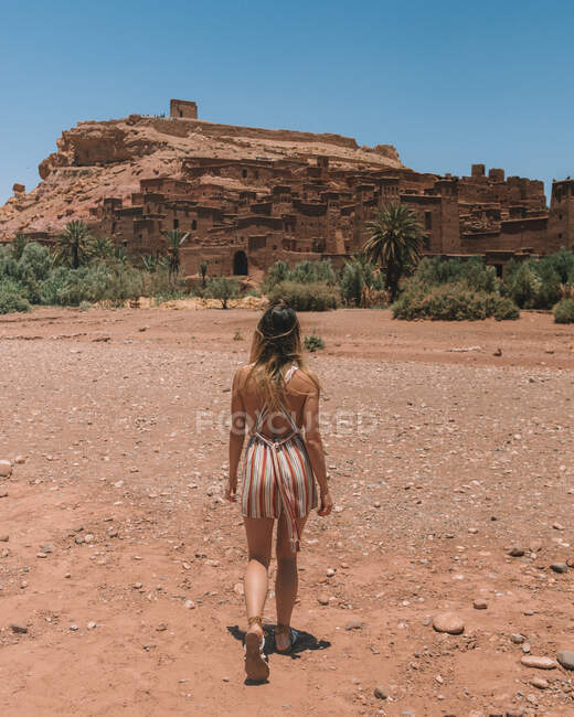 Back view of stylish woman walking on dry terrain of Moroccan desert against aged stone city in rock — Stock Photo