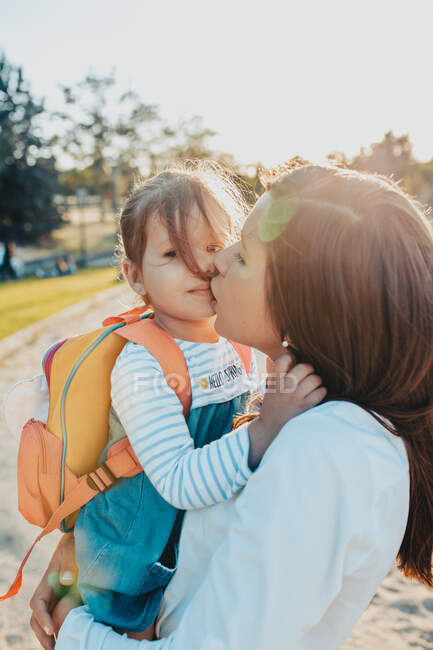 Young woman holding and kissing little girl with backpack while spending time in park together on sunny day — Stock Photo