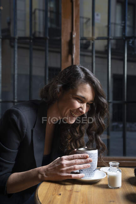 Vista laterale di bella donna con i capelli lunghi seduti al tavolo di legno dalla finestra e fare il tè — Foto stock