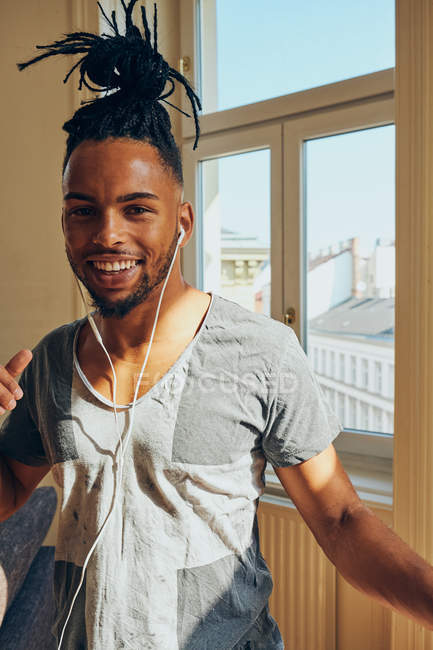 Smiling African American man with braids listening to music with earphones at home on window background — Stock Photo