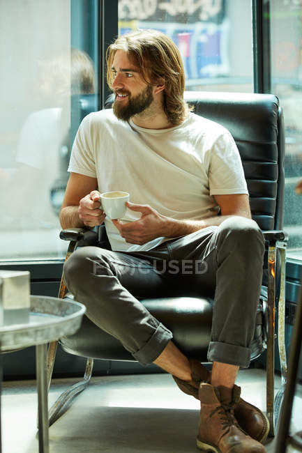 Молодий бородатий красивий чоловік сидить тримаючи чашку кави в руках і сміючись — стокове фото