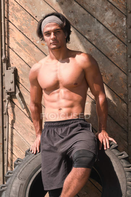 Muscular man posing in gym on tire in front on wooden wall — Fotografia de Stock