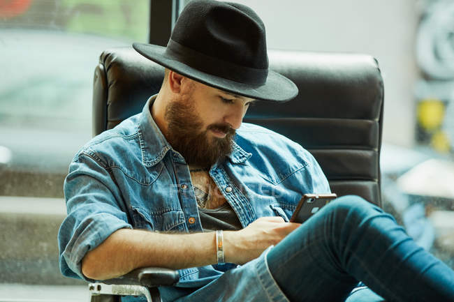 Молодий бородатий красивий чоловік у капелюсі та джинсовій сорочці задумливо переглядає мобільний телефон, сидячи в чорному кріслі — стокове фото