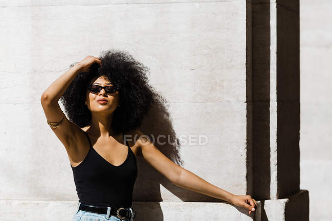 Молода етнічна жінка в джинсах і танк зверху спирається на стіну і посміхається на камеру на відкритому повітрі — стокове фото