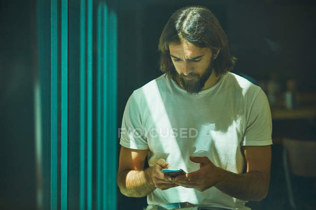 Молодий бородатий красивий чоловік спирається на стіну з мобільним телефоном в руках вдумливо смс — стокове фото