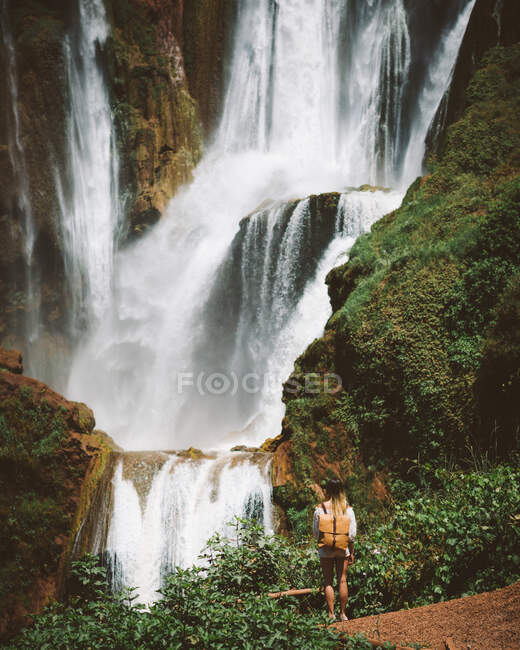 Visão traseira da mulher em pé em terreno deserto com poderosa cachoeira majestosa em falésias verdes, Marrocos — Fotografia de Stock