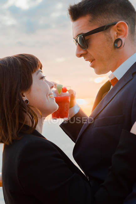 Side view of young attractive couple drinking red beverage with straws from one glass on background of sunset sea — Stock Photo