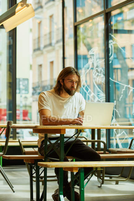 Junger bärtiger schöner Mann sitzt draußen im Café und arbeitet mit Laptop auf dem Tisch — Stockfoto