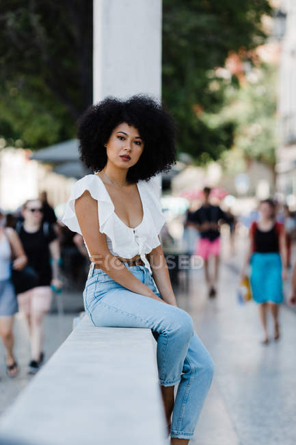 Young African American woman in jeans and crop top relaxing on stone railing and looking at camera outdoors — Stock Photo