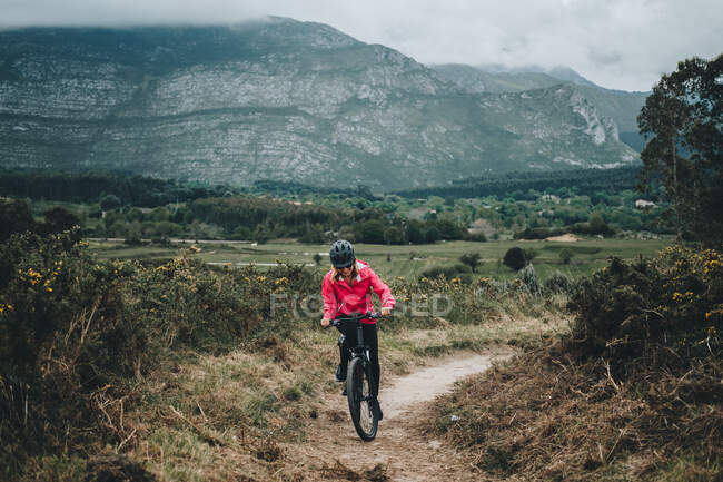 Active woman in helmet and red jacket riding bicycle on countryside road on nature background — Stock Photo