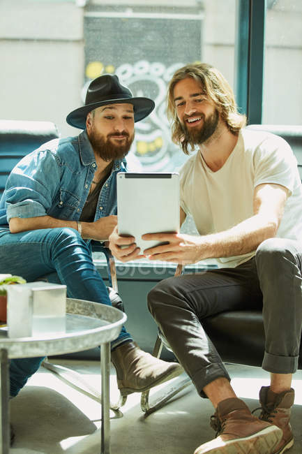 Красивый мужчина в черной шляпе сидит и смотрит на экран планшетного компьютера с другом в кафе — стоковое фото