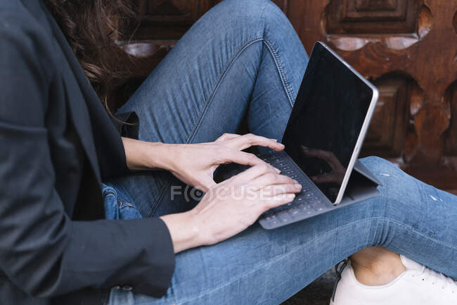 Seitenansicht einer nicht wiedererkennbaren Frau, die ein digitales Tablet benutzt, während sie sich auf der Schwelle einer alten Holztür entspannt — Stockfoto