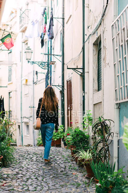 Rückansicht einer lässigen Frau mit Handtasche, die auf der gepflasterten schmalen Straße der Altstadt mit Blumentöpfen an den Seiten spaziert — Stockfoto