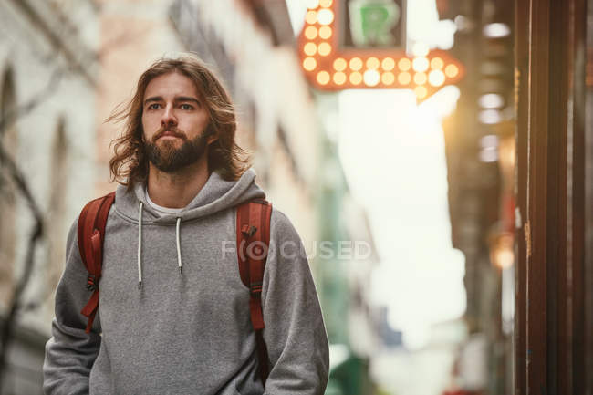 Junger bärtiger, gutaussehender Mann in grauem Kapuzenpulli mit Rucksack geht ernsthaft nach draußen — Stockfoto