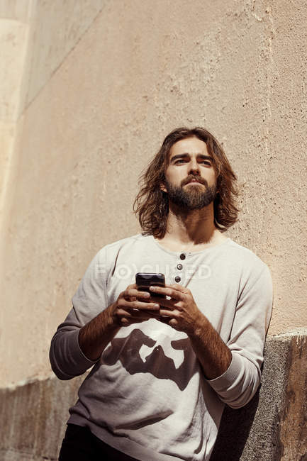 Молодий бородатий красивий чоловік у бежевому светрі спирається на стіну і тримає мобільний телефон в руках вдумливо дивлячись вздовж — стокове фото