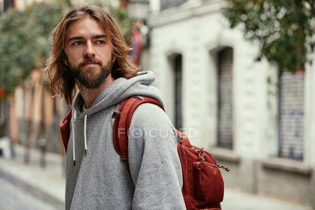 Junger bärtiger, gutaussehender Mann in grauem Kapuzenpulli mit Rucksack geht ernsthaft nach draußen — Stockfoto