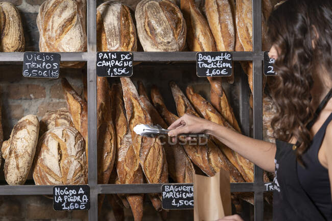 Unrecognizable woman in black apron putting French baguette in paper bag by metal thongs in bakery - foto de stock