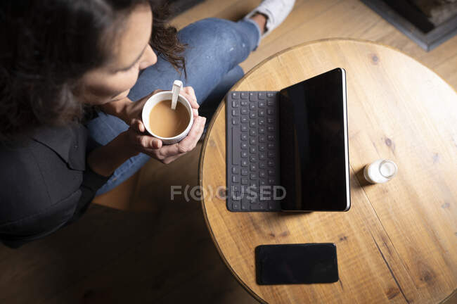 Зверху - невпізнавана жінка, яка сидить за столом з чашкою чаю і користується цифровим планшетом і смартфоном. — стокове фото