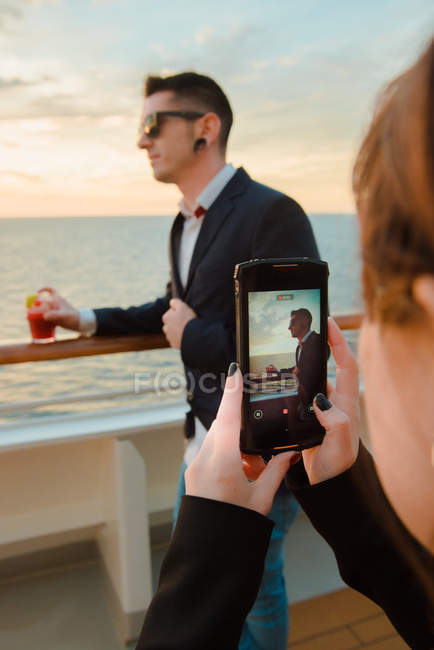 Молодой красавец в черных солнцезащитных очках с бокалом красного напитка стоит на палубе корабля, а женщина солнечным вечером фотографирует на мобильный телефон — стоковое фото