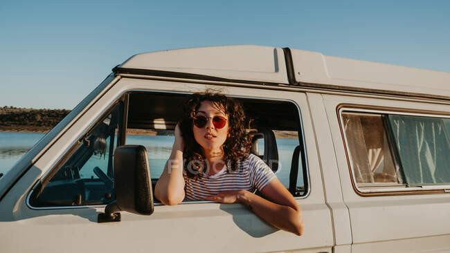 Bastante joven morena sonriendo y mirando hacia otro lado mientras se sienta dentro de un coche vintage sobre un fondo borroso de la naturaleza - foto de stock