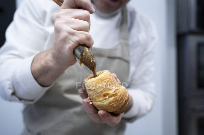 Homem de colheita em avental e uniforme branco recheando croissant fresco com creme de chocolate dentro de casa — Fotografia de Stock