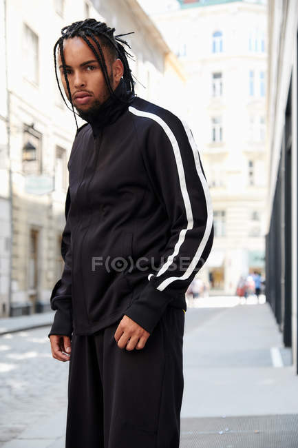 Молодой этнический человек с плетеными волосами в черном спортивном костюме смотрит в камеру на городском фоне — городские, Прическа - Stock Photo