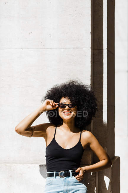 Young ethnic woman in jeans and tank top leaning on wall and smiling at camera outdoors — Stock Photo
