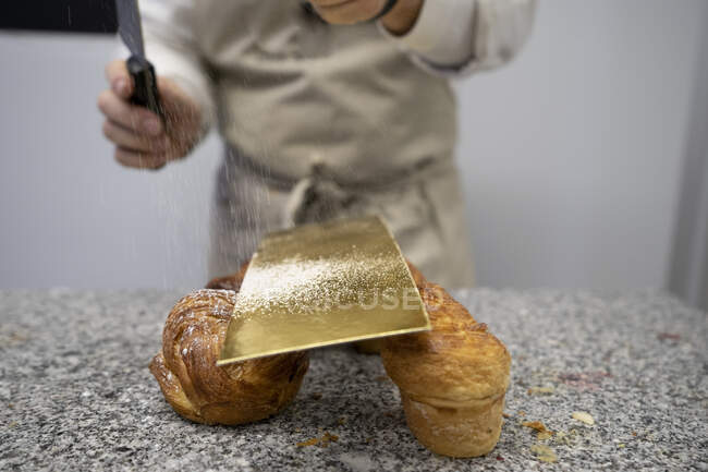 Кукурузник просеивает сахар на свежей выпечке, расположенной на каменном столе с помощью ножа и золотой фольги — стоковое фото