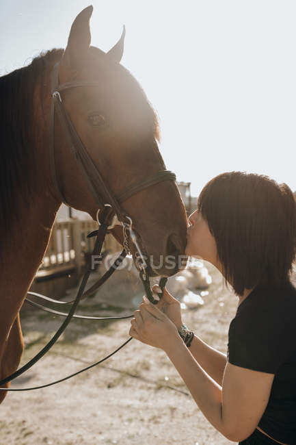 Молодая женщина с закрытыми глазами целует лошадь, проводя время на ранчо в солнечный день — стоковое фото