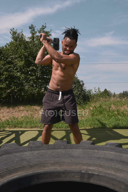 Strong shirtless African American guy hitting tire with heavy hammer while working out in outdoor gym — Fotografia de Stock