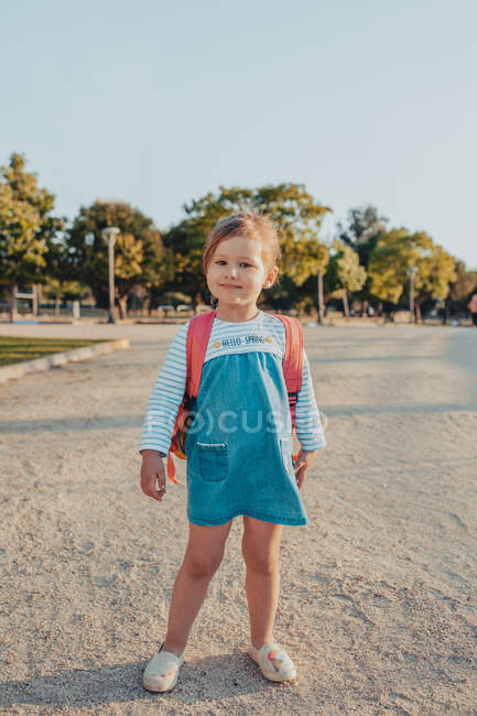 Cute little girl with backpack smiling and looking at camera while standing on sandy ground in park — Stock Photo