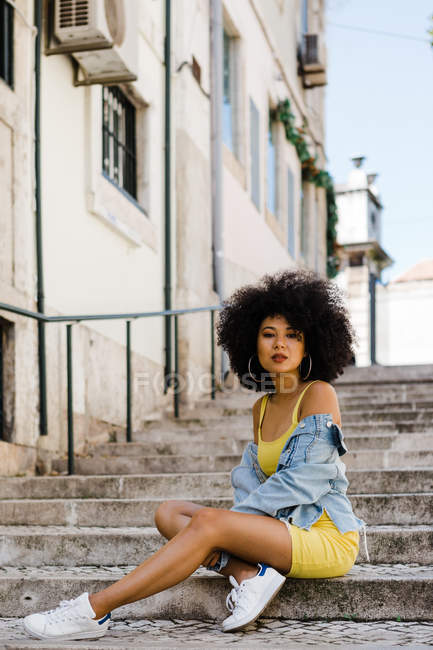 Stylish African American woman in yellow suit and denim jacket sitting and looking at camera on urban background — Stock Photo