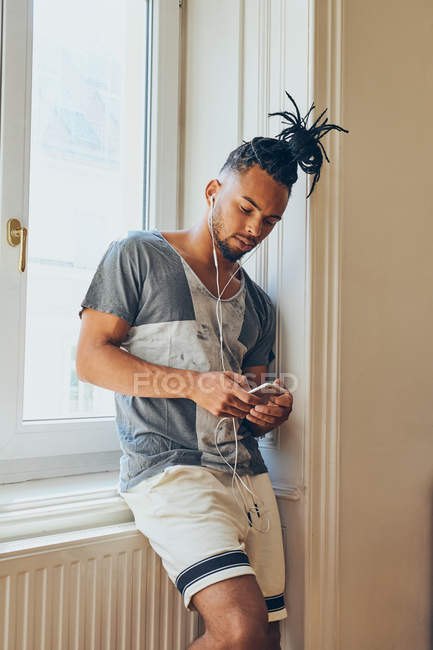 Young African American man with creative hairstyle leaning on windowsill at home using mobile phone and listening to music with earphones — Stock Photo