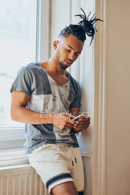 Молодий афроамериканець людина з творчою зачіскою спираючись на підвіконня в домашніх умовах за допомогою мобільного телефону — стокове фото