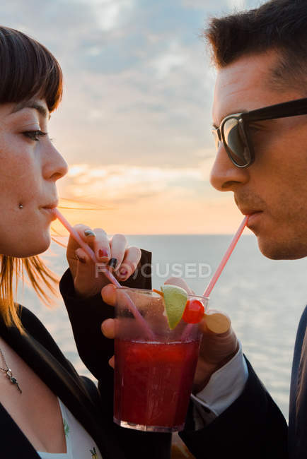 Вид сбоку молодой привлекательной пары, пьющей красный напиток с соломинкой из одного бокала на фоне закатного моря — стоковое фото