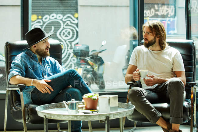 Beau homme au chapeau noir assis et profitant de la conversation avec un ami tenant une tasse de café dans un café — Photo de stock