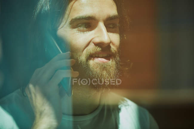 Joven barbudo guapo hombre en la cafetería detrás de la ventana hablando por teléfono - foto de stock