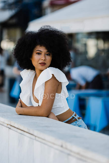 Young African American woman in jeans and crop top leaning on stone railing and looking at camera outdoors — Stock Photo