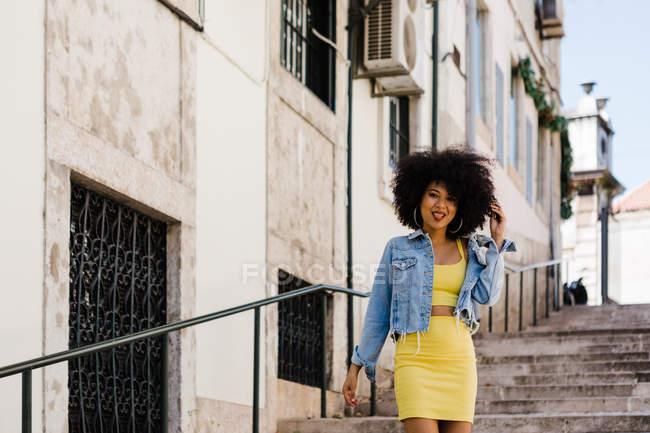 Улыбающаяся афроамериканка в жёлтом костюме, стоящая на лестнице и смотрящая в камеру на городском фоне — стоковое фото