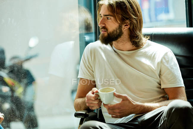 Young pensive bearded handsome man sitting holding cup of coffee in hands — Stock Photo