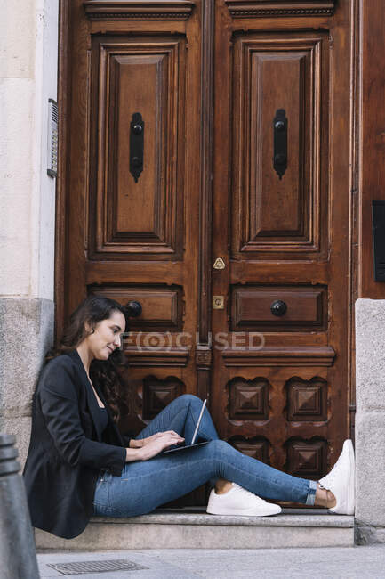 Vista lateral de la hermosa mujer usando tableta digital mientras se relaja en el umbral de la puerta de madera vieja - foto de stock