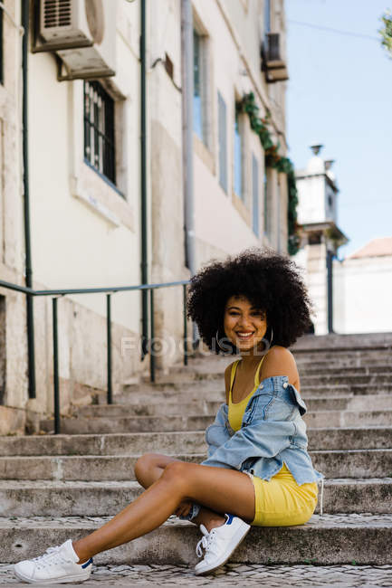 Femme afro-américaine souriante en costume jaune et veste en denim assis et regardant la caméra sur fond urbain — Photo de stock